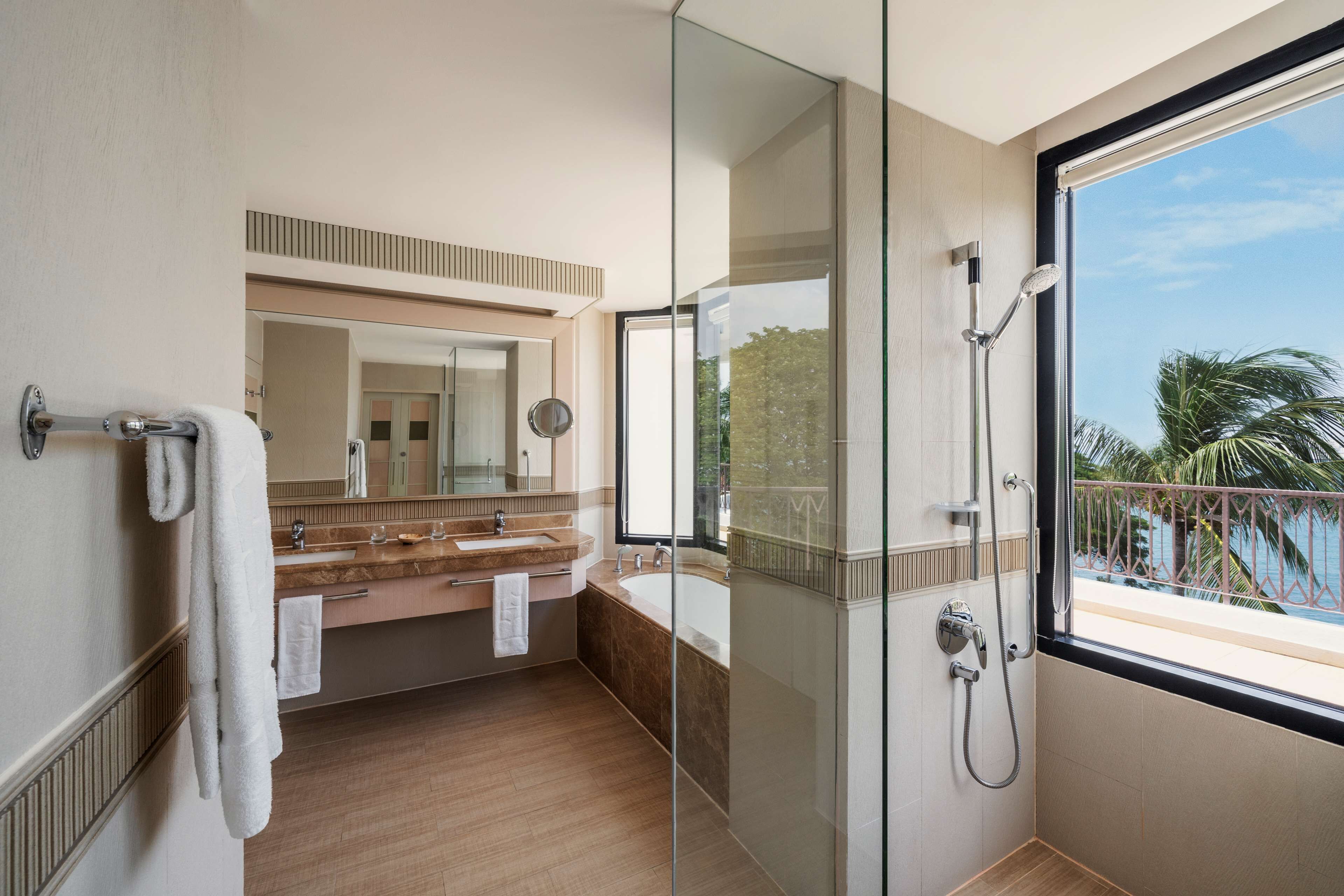 Un baño moderno con cabina de ducha de vidrio, un espejo grande y vista al mar desde un balcón.