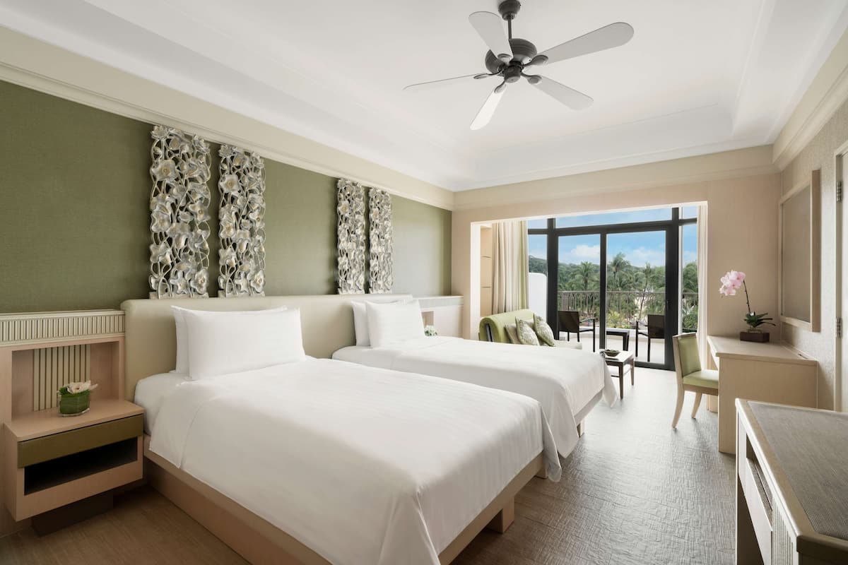 A hotel room with two beds, a balcony with a view, a desk, and a ceiling fan.