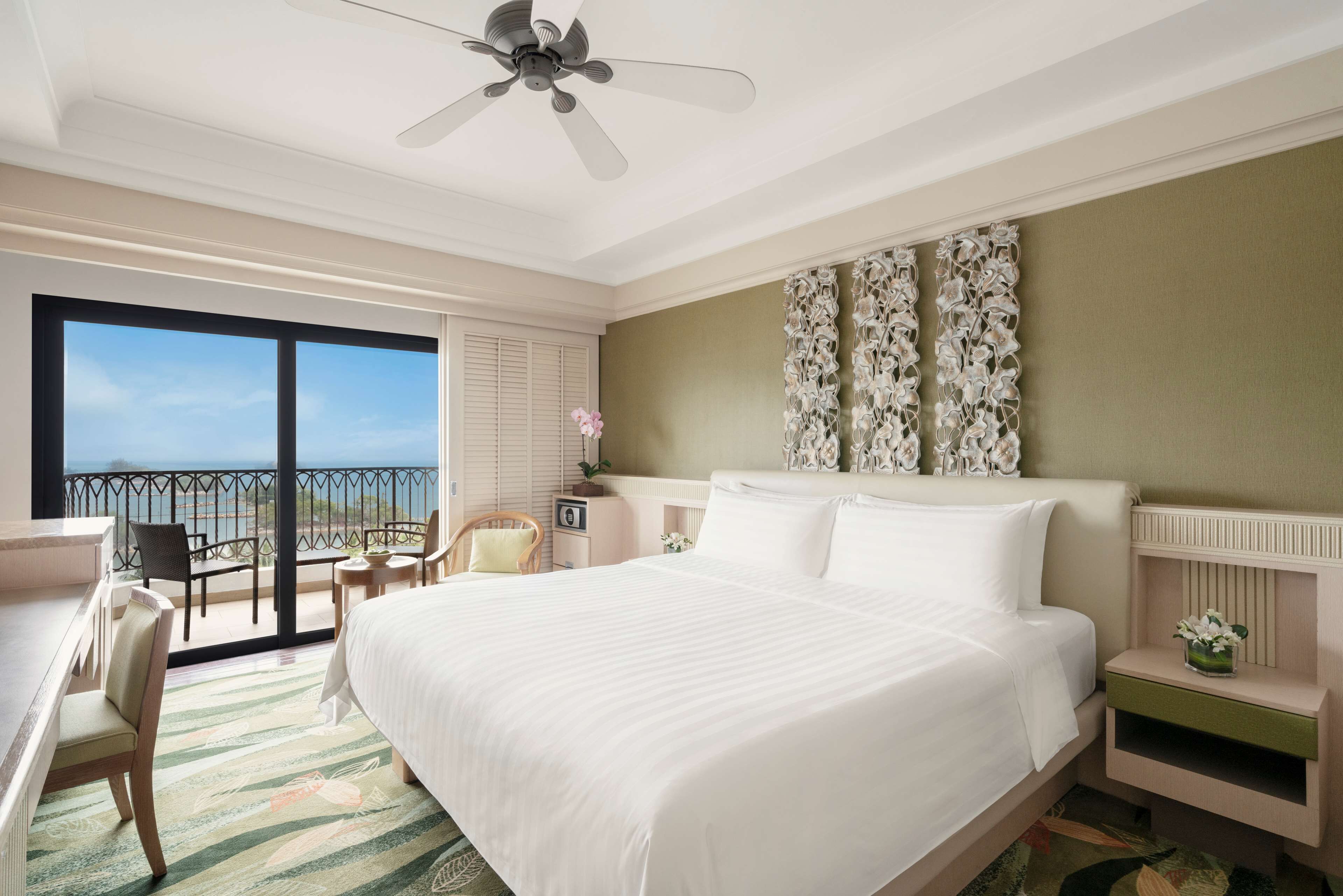 A hotel room with a large bed, a desk, a chair, a small table, a nightstand, and a balcony with a view of the ocean.