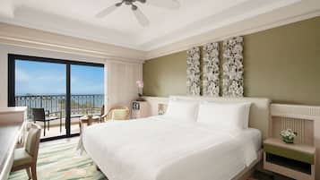 A hotel room with a large bed, a desk, a chair, a small table, a nightstand, and a balcony with a view of the ocean.