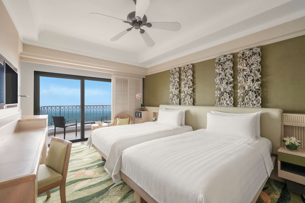 A hotel room with two beds, a desk, a chair, a balcony with a view, and a ceiling fan.
