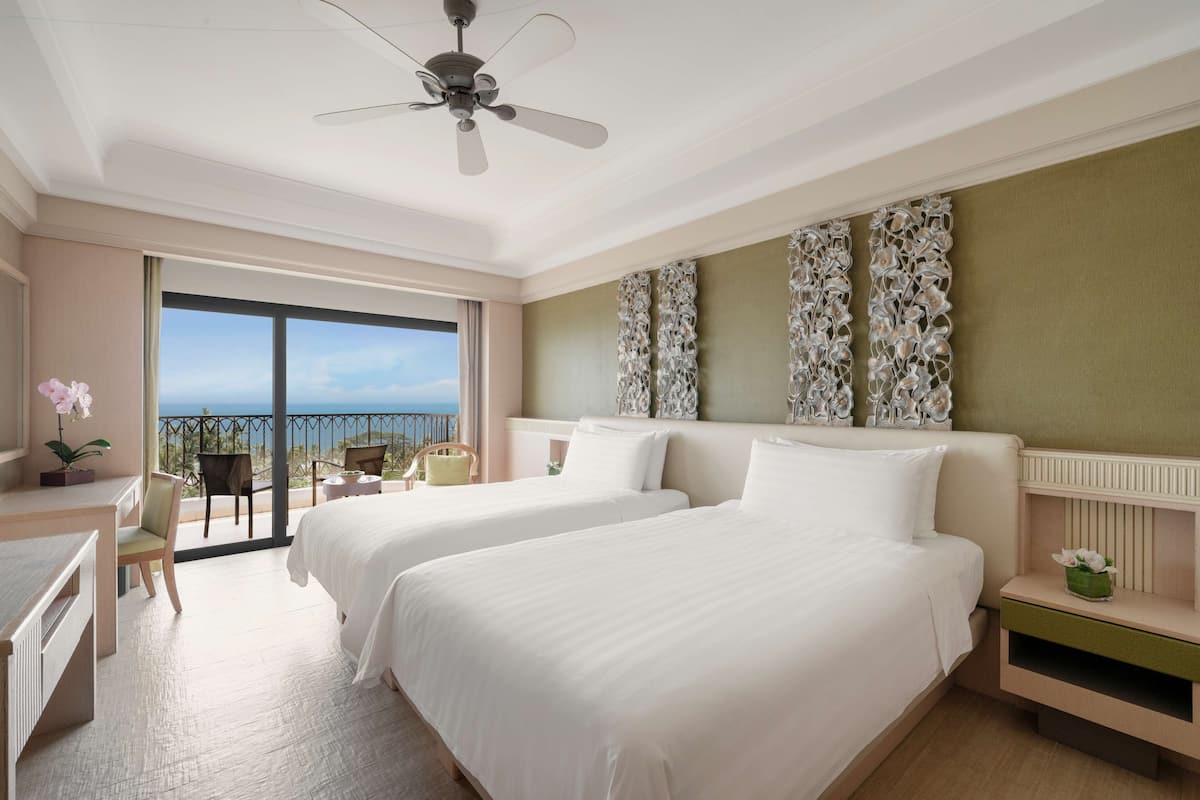 A hotel room with two beds, a desk, a chair, and a balcony with a view of the sea.