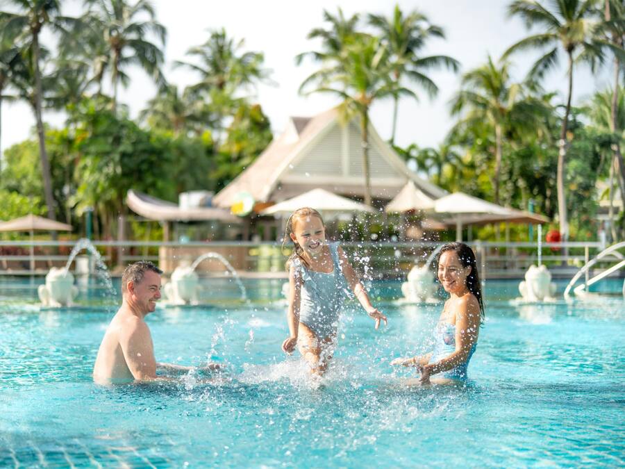 A swimming pool with water fountains, palm trees, and umbrellas in the background.