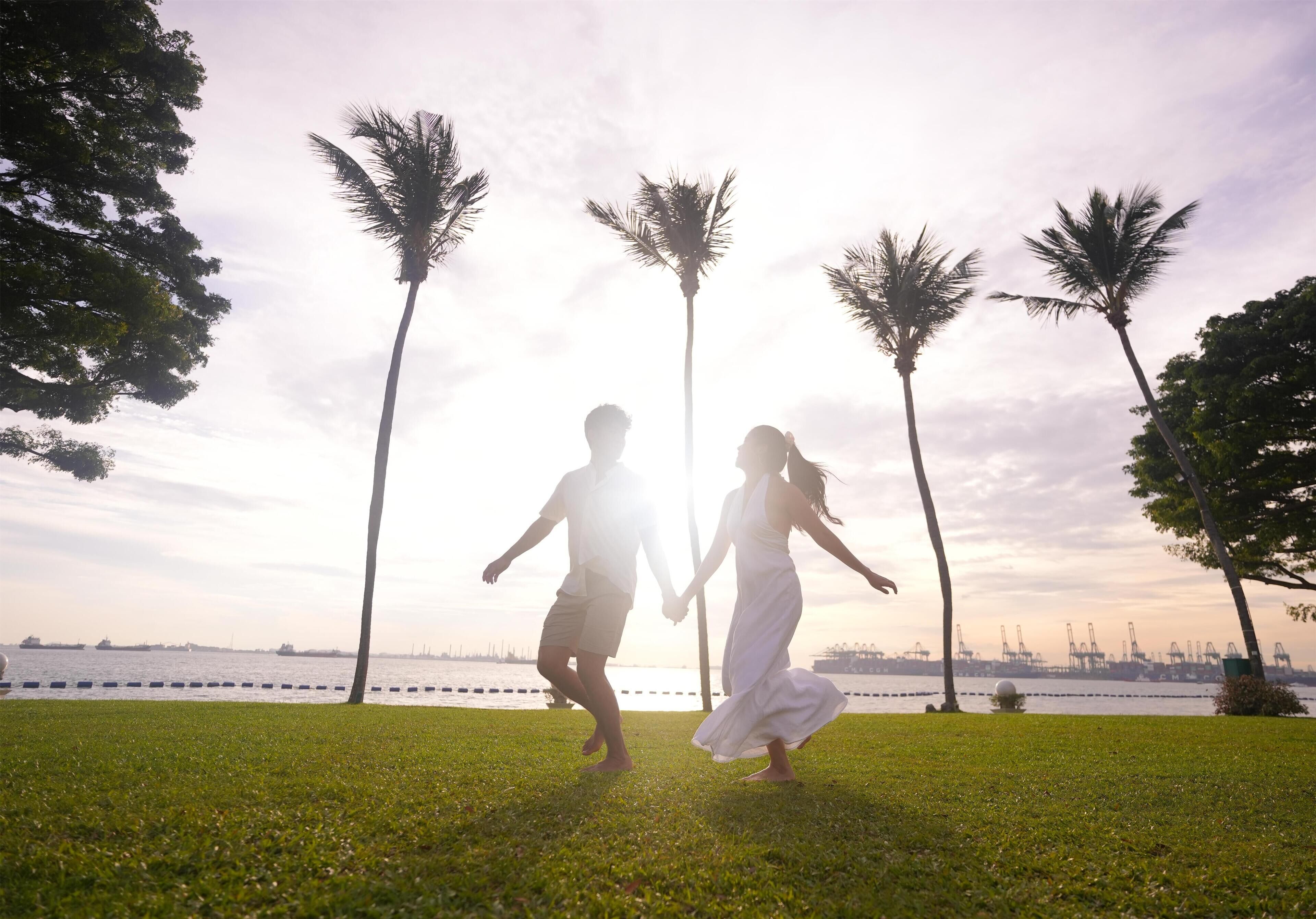 A couple holding hands on a grassy area with palm trees and a body of water in the background.