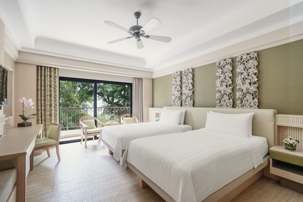 A hotel room with two beds, a desk, a chair, a ceiling fan, and a balcony with a view of trees.