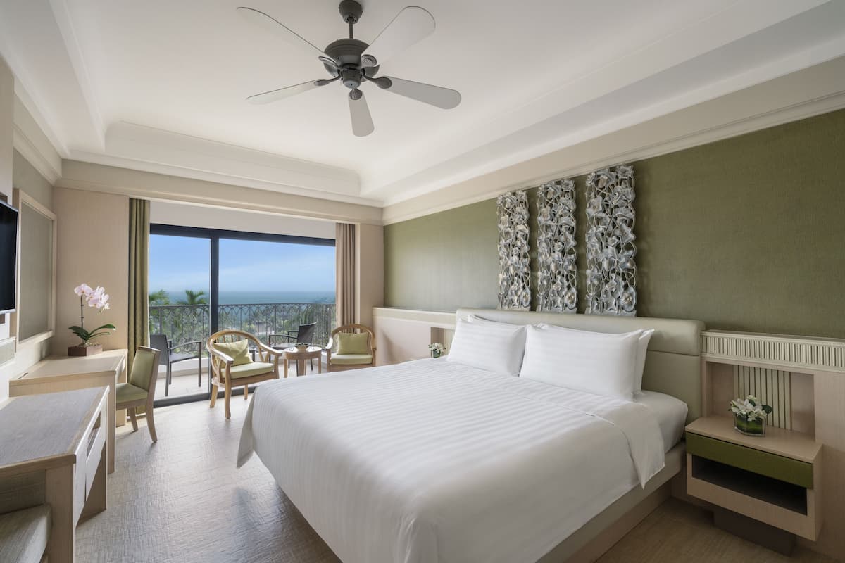 A hotel room with a large bed, a desk, a chair, a small table, a ceiling fan, and a balcony with a view.