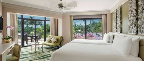 A hotel room with a large bed, a desk, a chair, a small table, and a balcony with a view of palm trees and flowers.