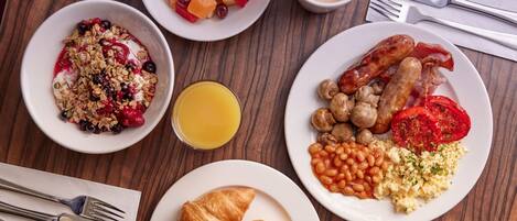 Una mesa de desayuno con diversos platos, como cereal, fruta, salchichas, frijoles y huevos.