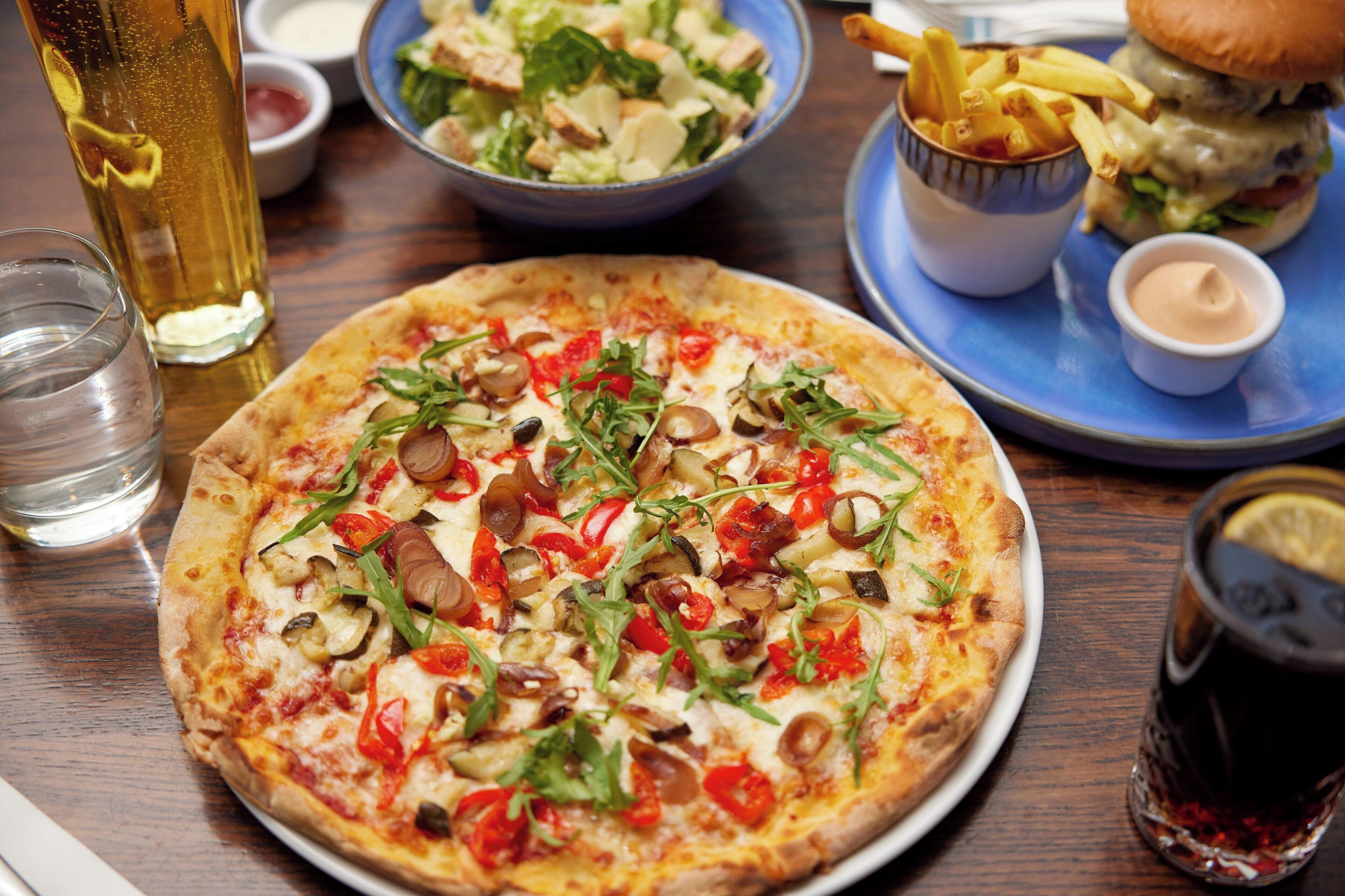 A pizza with various toppings, a salad, fries, a burger, and a drink on a table.