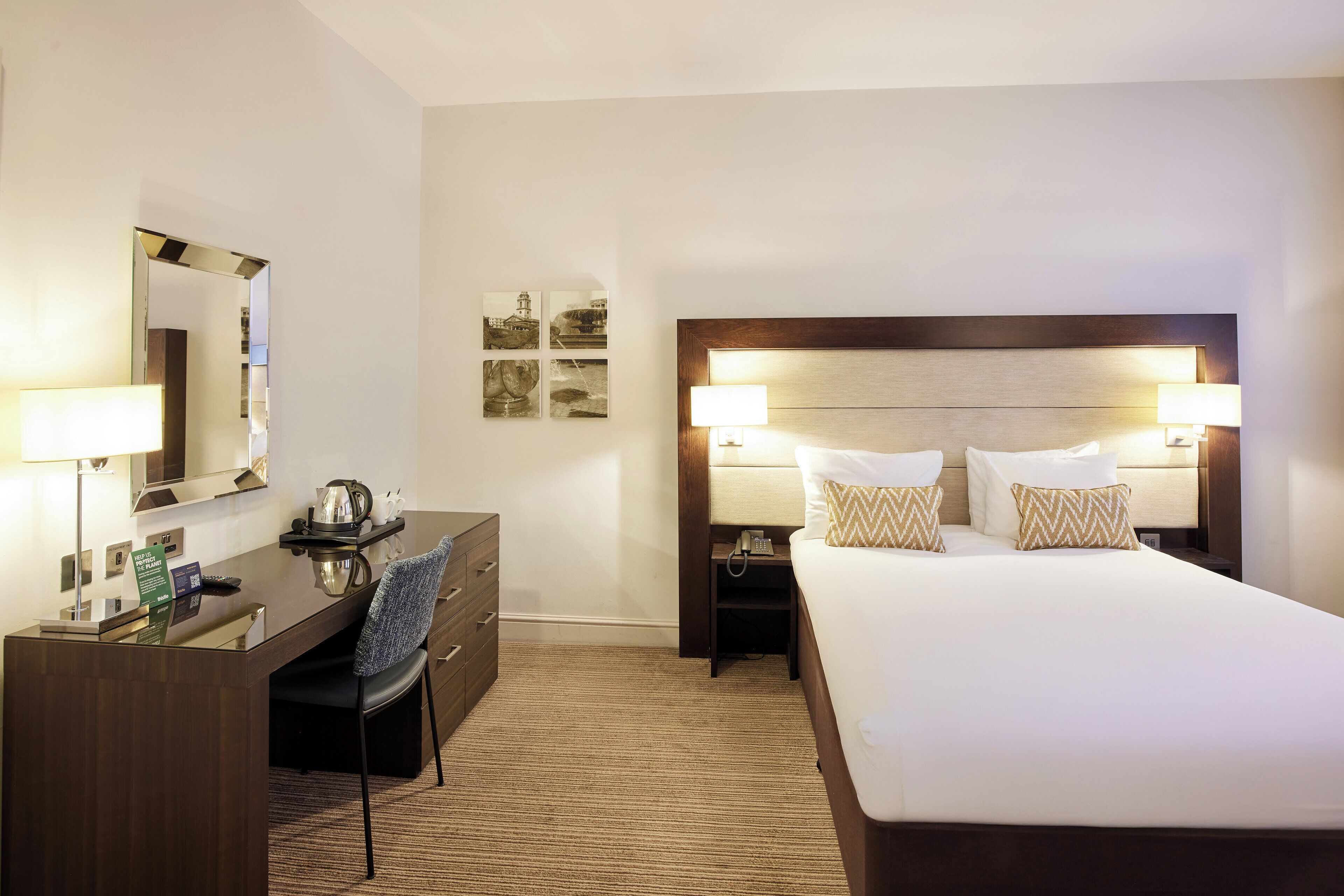 A hotel room with a large bed, two bedside tables with lamps, a desk with a chair, a mirror, and a small coffee table.