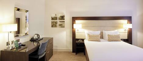 A hotel room with a large bed, two bedside tables with lamps, a desk with a chair, a mirror, and a small coffee table.