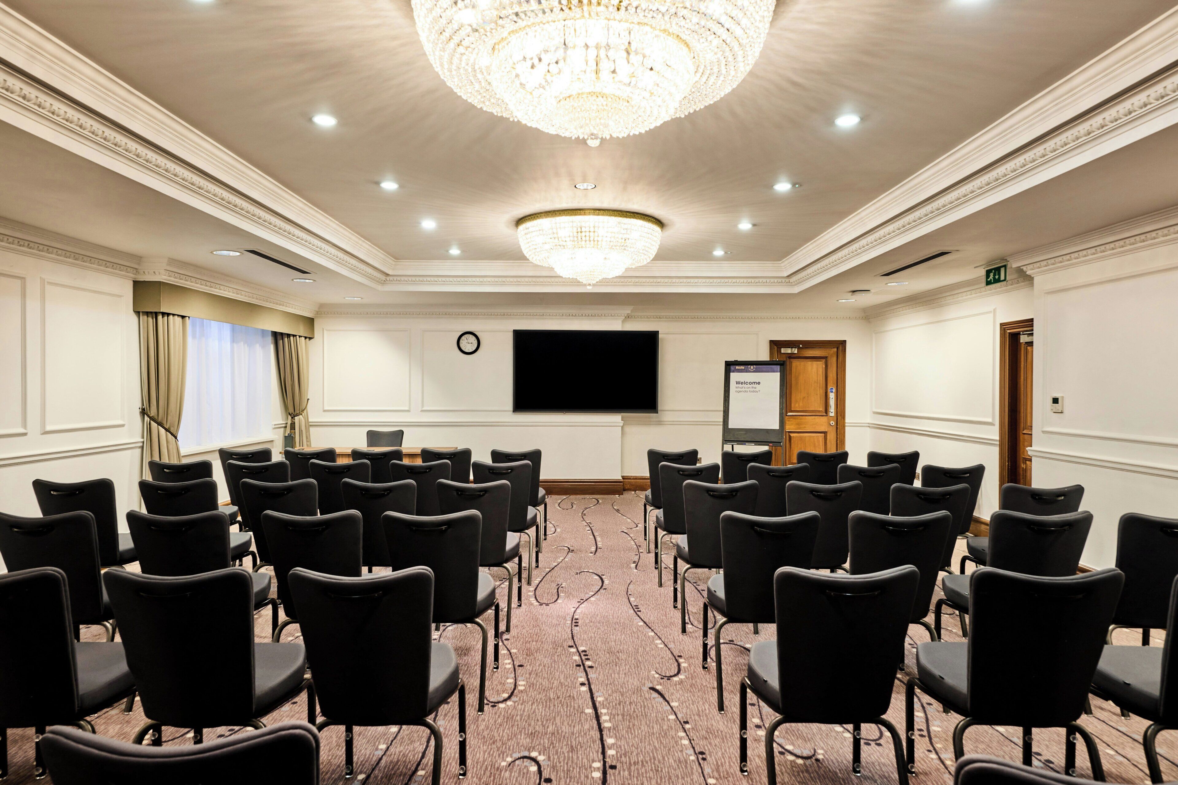 Une salle de conférence avec des rangées de chaises, un grand écran et un lustre.