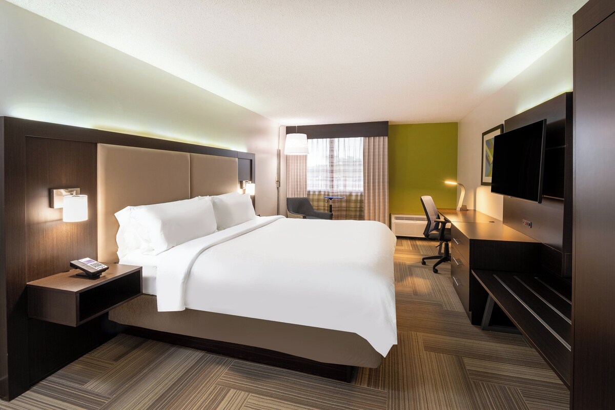 A hotel room with a large bed, a desk with a chair, a flat-screen TV, and a window with curtains.