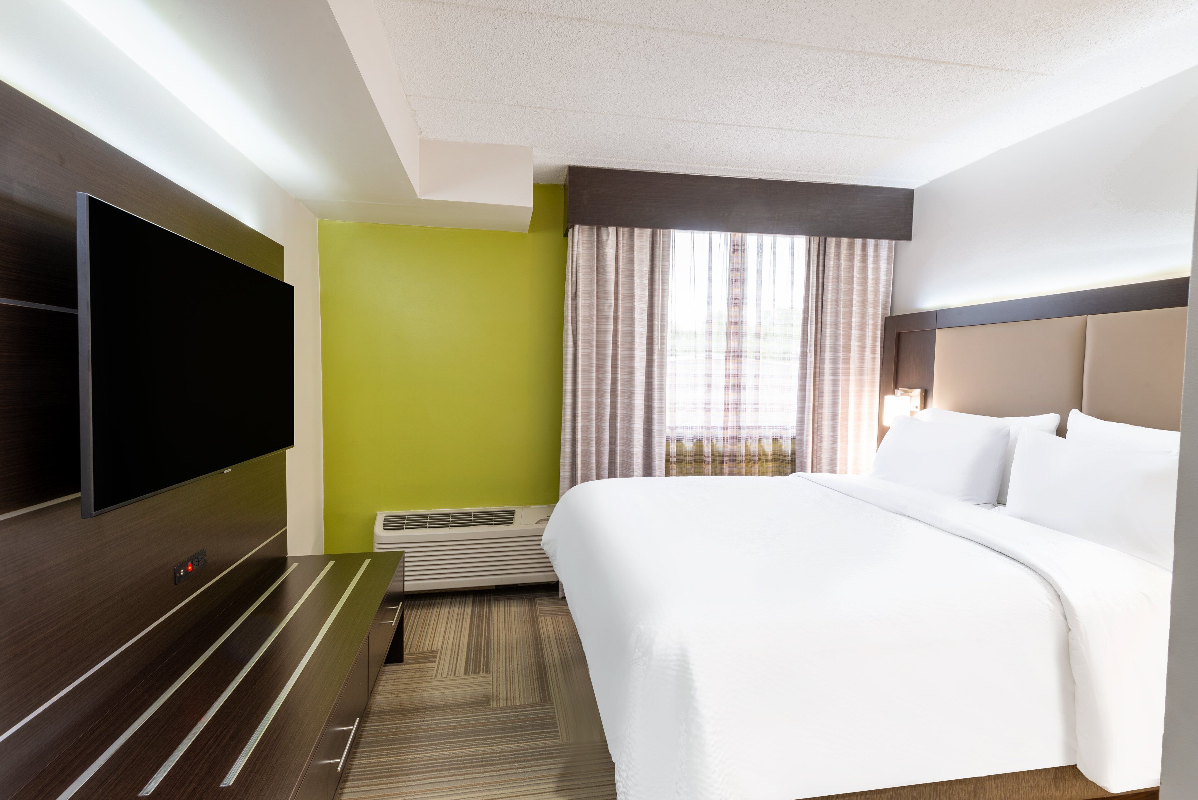 A hotel room with a large bed, a flat-screen TV mounted on the wall, a small table, and a window with curtains.