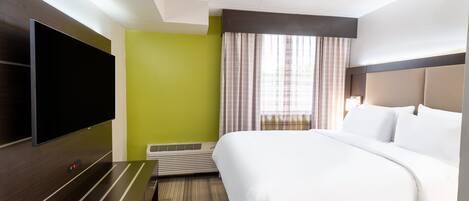 A hotel room with a large bed, a flat-screen TV mounted on the wall, a small table, and a window with curtains.