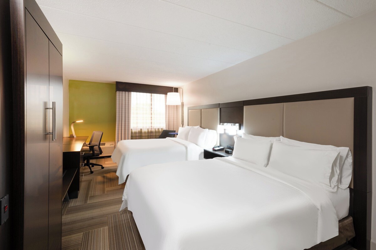 A hotel room with two beds, a desk, a chair, and a lamp.