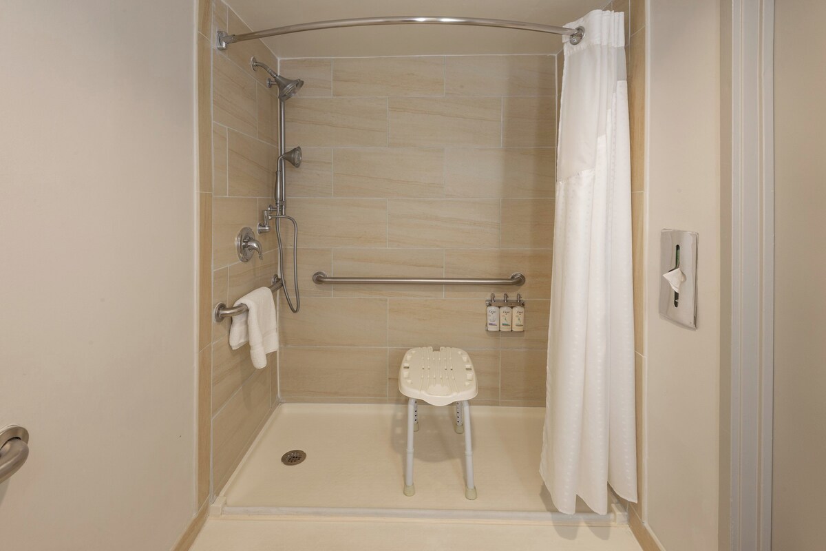 A walk-in shower with a white shower chair, grab bars, and a handheld showerhead.