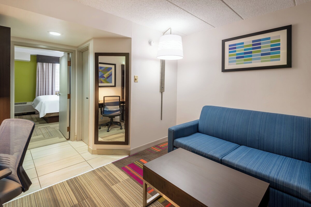 A hotel room with a blue sofa, a wooden coffee table, a chair, a desk, and a framed abstract art piece on the wall.