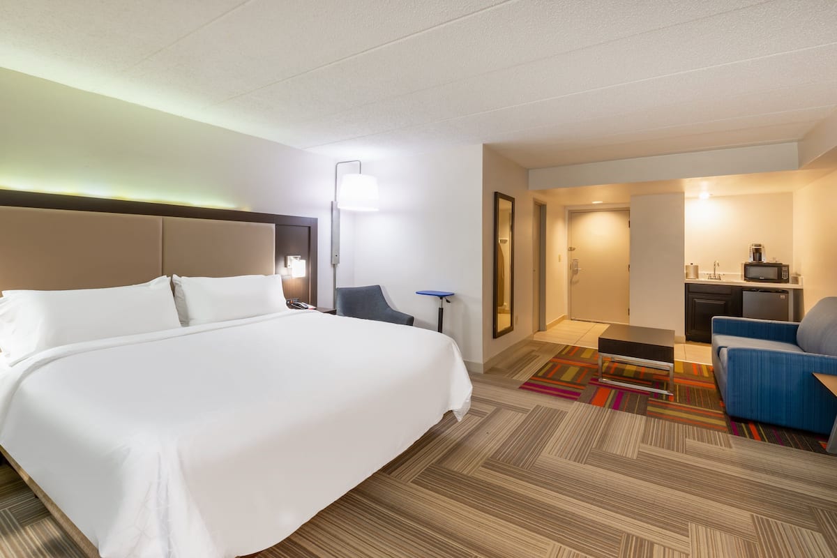 A hotel room with a large bed, a desk, a chair, a television, and a kitchenette.