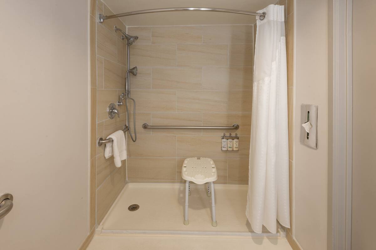 A walk-in shower with a white shower chair, grab bars, and a handheld showerhead.