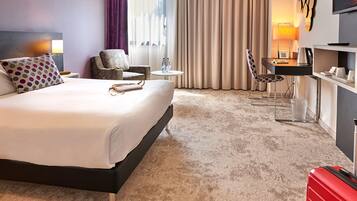 A hotel room with a bed, a chair, a desk, a TV, and a red suitcase.