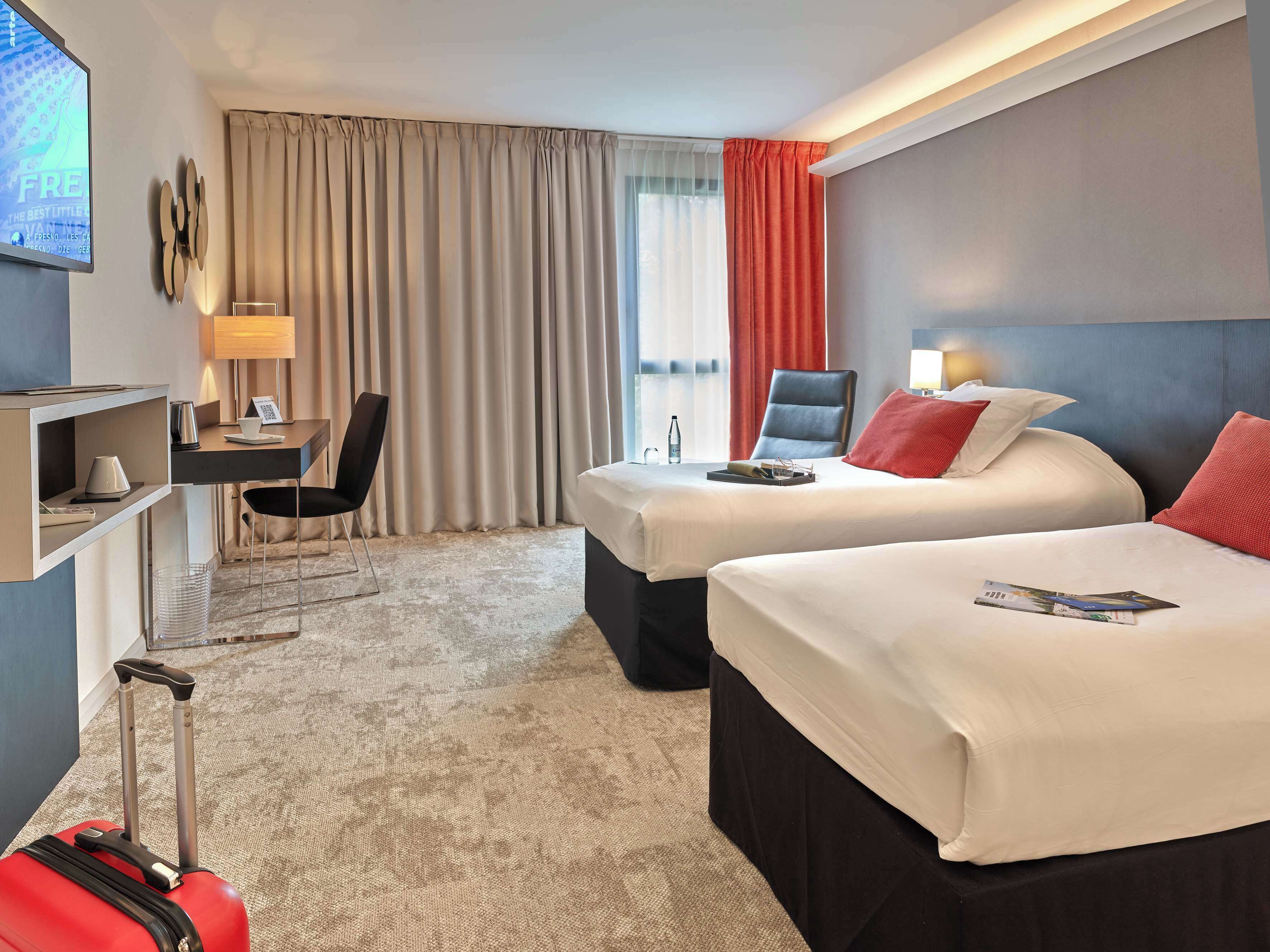 A hotel room with two beds, a desk, a chair, a TV, and a suitcase.