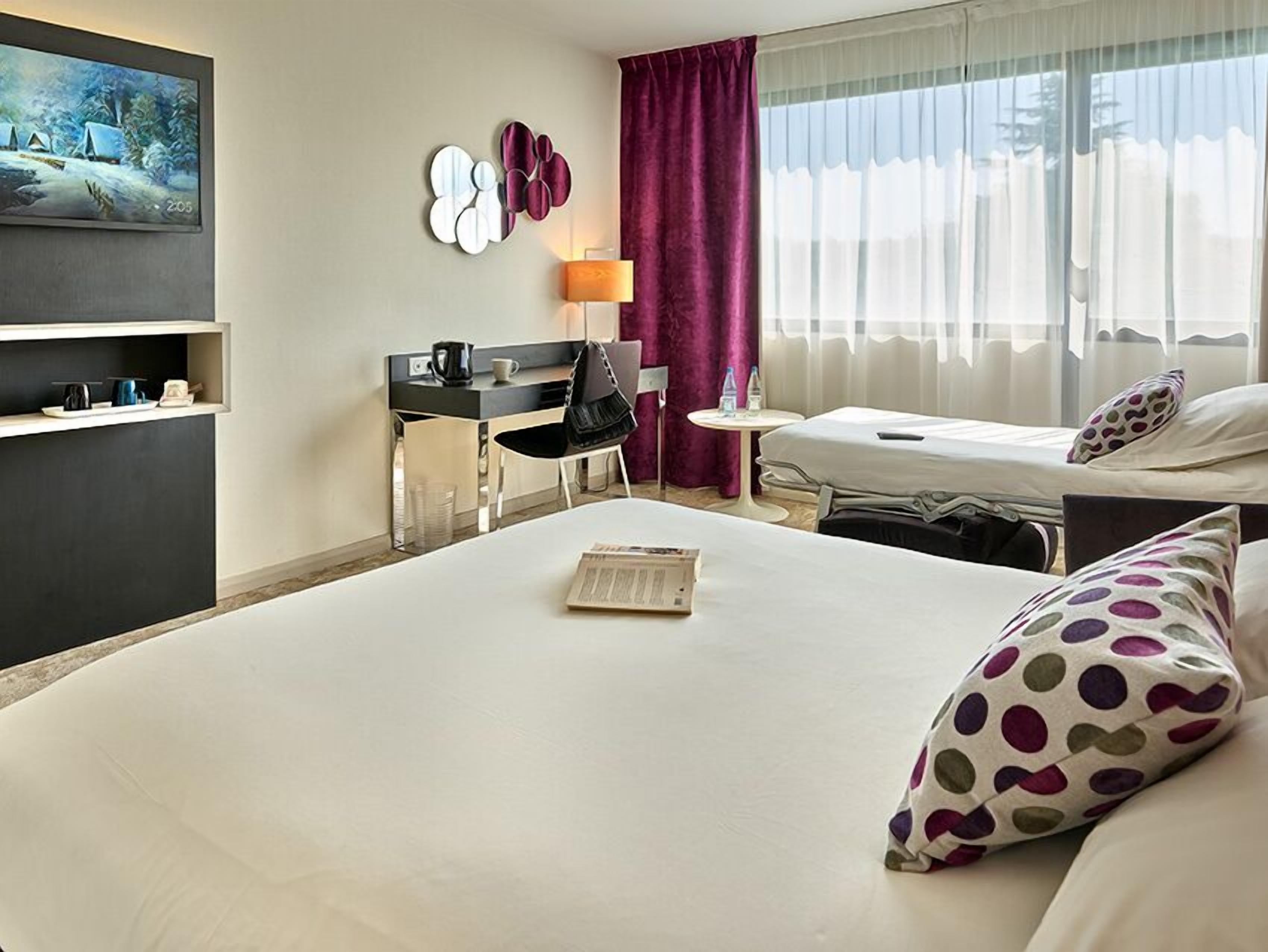Habitación de hotel con cama, escritorio, silla, mesita, televisor y cortinas.