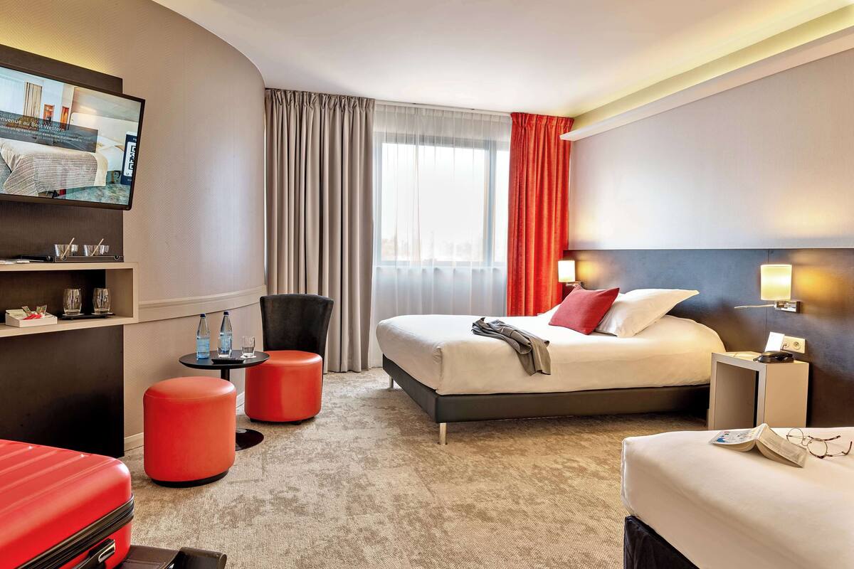 A hotel room with a bed, a TV, a desk, and a red suitcase.