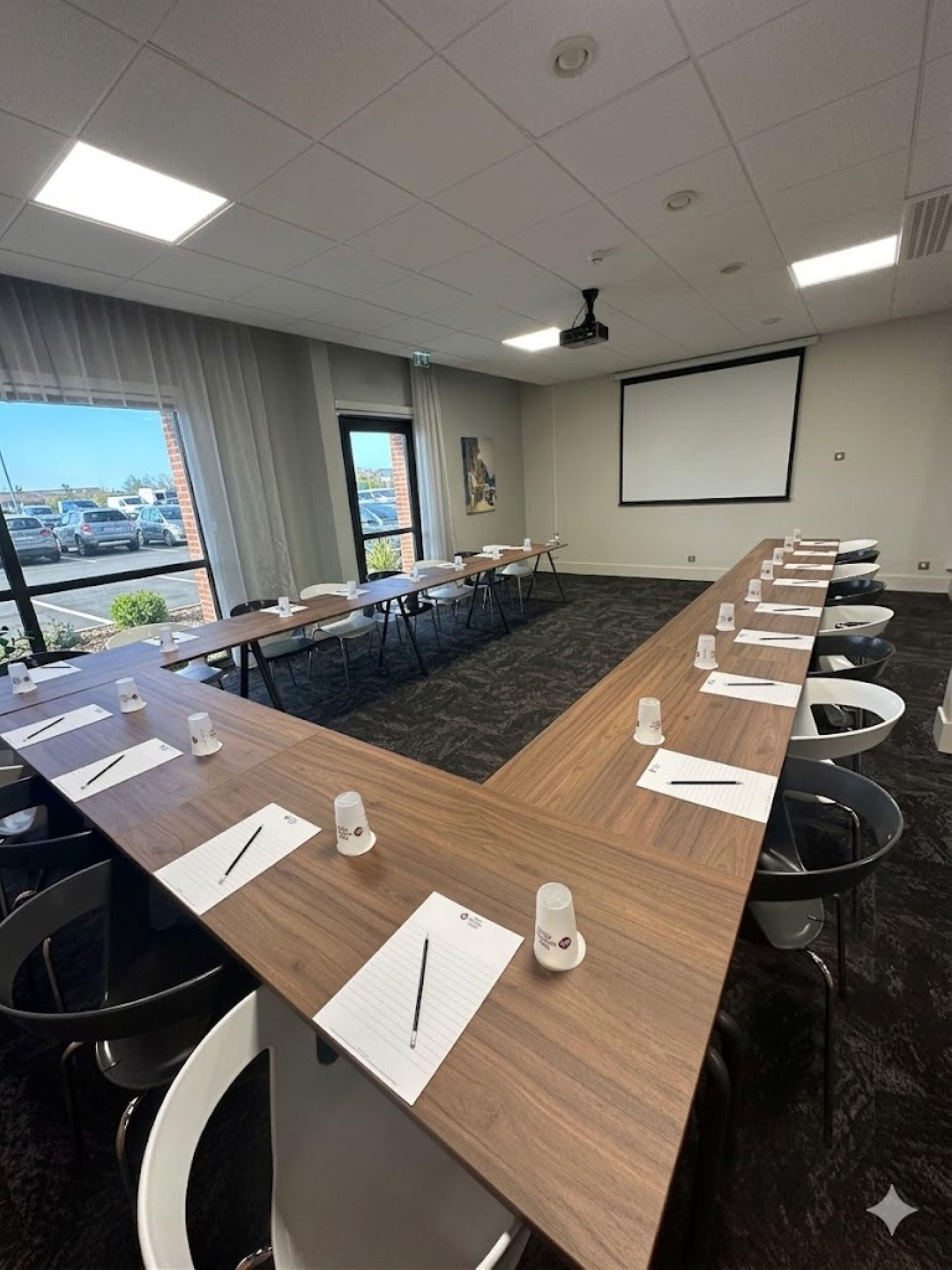 A conference room with a long table, chairs, a projector screen, and a view of a parking lot.