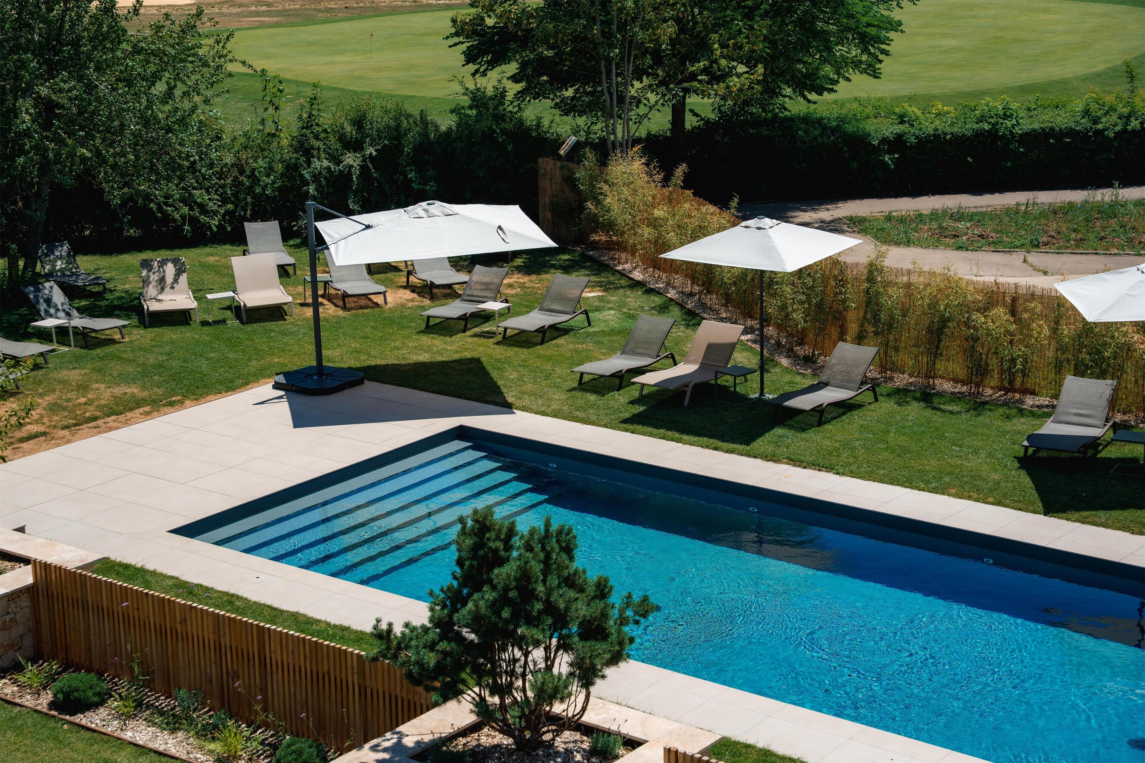A swimming pool with sun loungers and umbrellas, surrounded by a wooden fence and greenery.