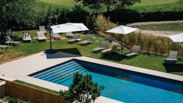 A swimming pool with sun loungers and umbrellas, surrounded by a wooden fence and greenery.