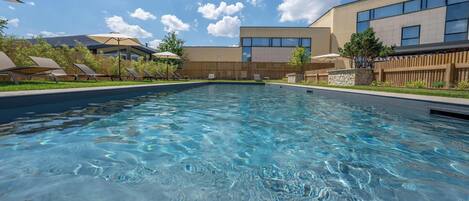 A clear swimming pool with lounge chairs and umbrellas, surrounded by a wooden fence and a building with large windows.