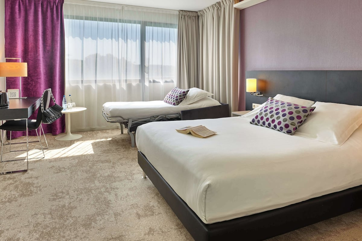 Quarto de hotel com uma cama grande, uma cama menor, uma escrivaninha com cadeira e vista para o exterior pela janela.