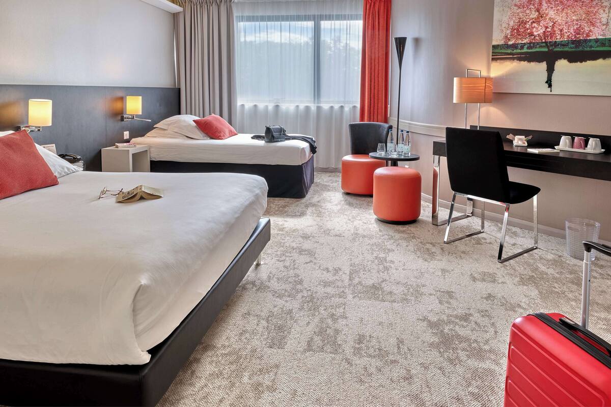 A hotel room with two beds, a desk, a chair, and a red suitcase.