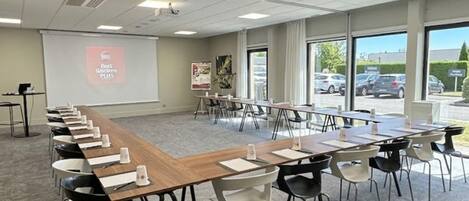 Ein Konferenzraum mit langem Tisch, Stühlen, einer Projektionsleinwand und großen Fenstern.