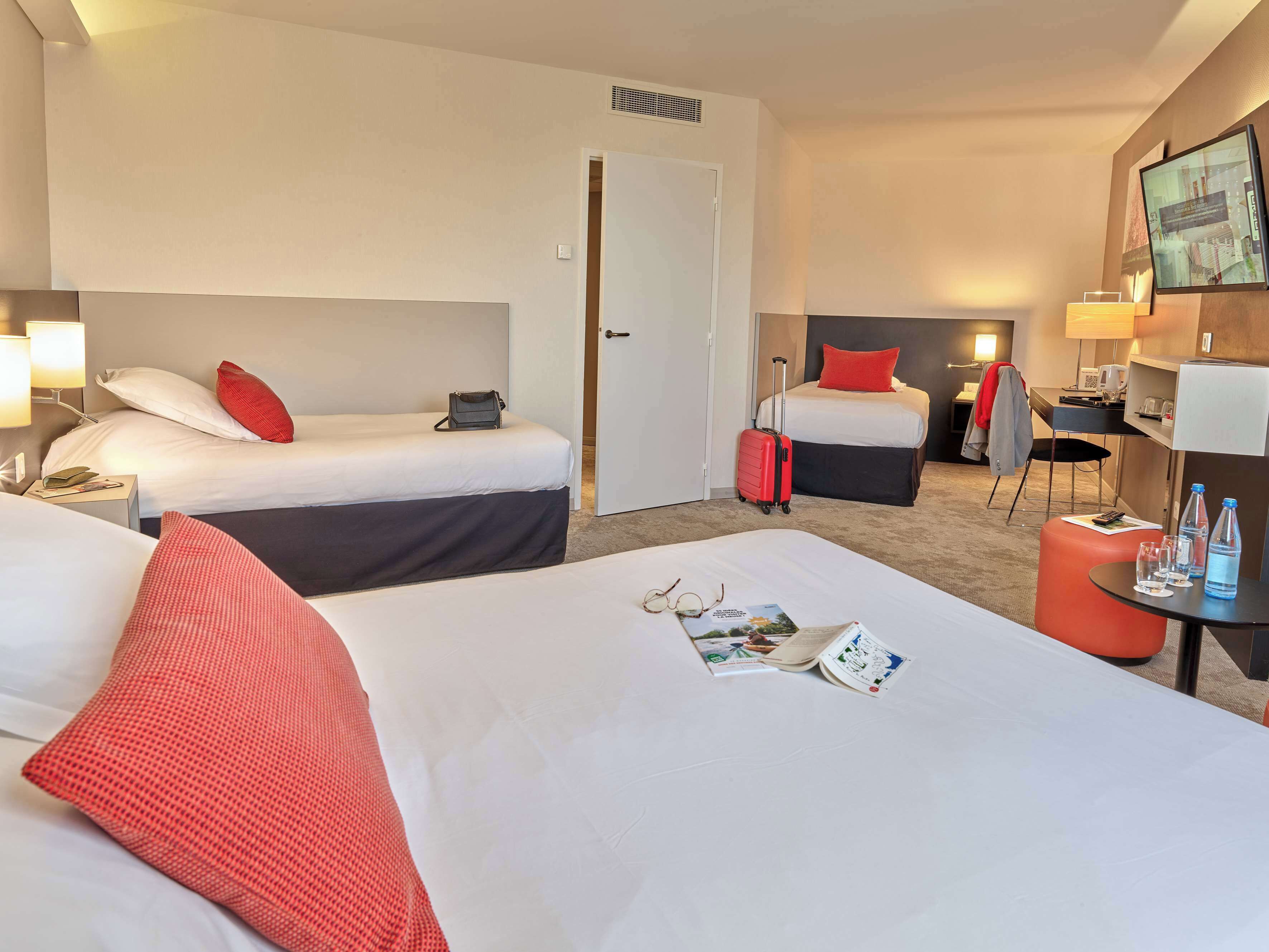 A hotel room with two beds, a desk, a chair, a TV, and a small table with glasses and a water bottle.