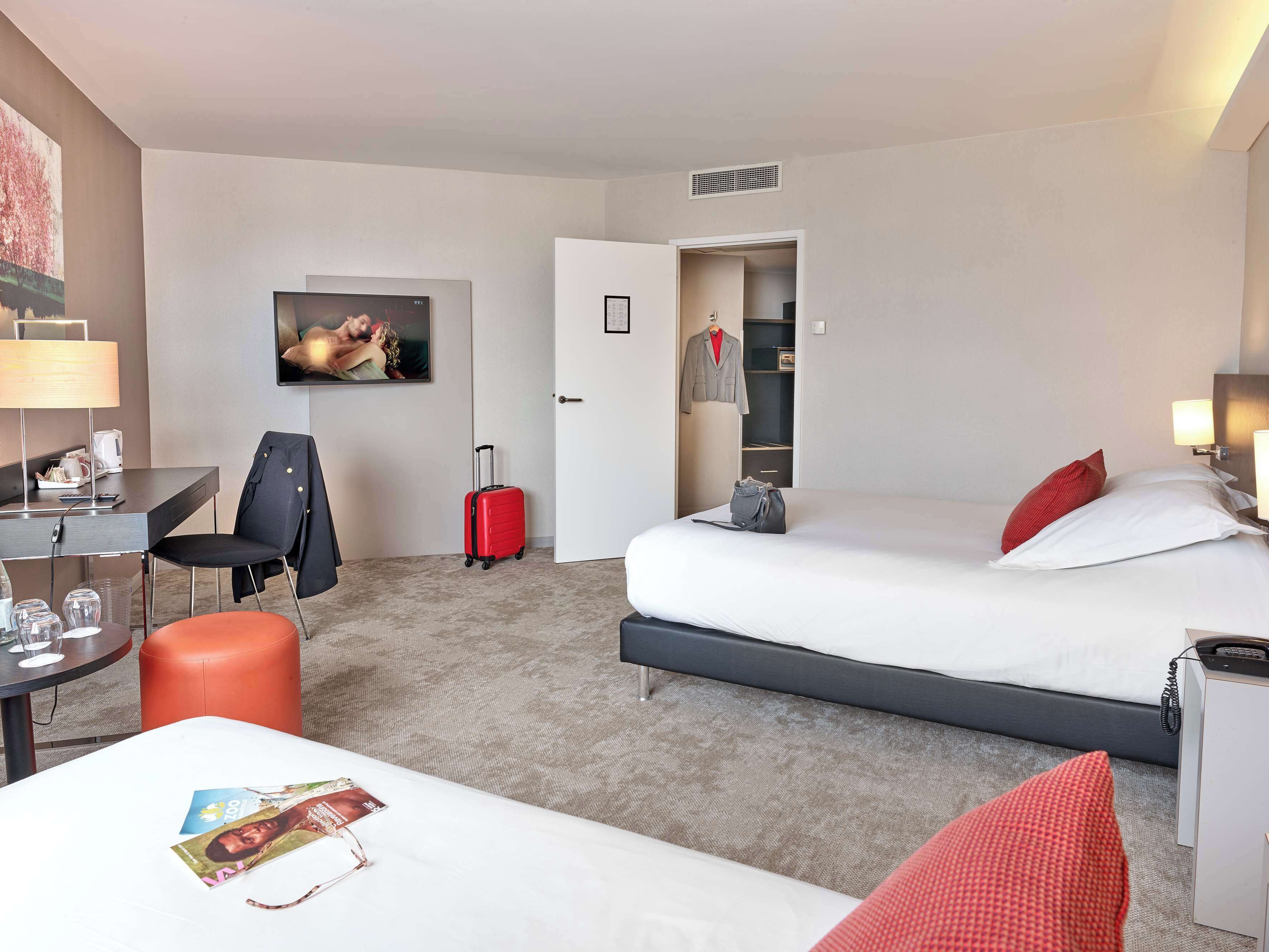 A hotel room with a bed, a desk, a chair, a red suitcase, a TV on the wall, and a wardrobe.