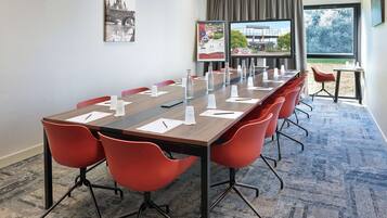 Een vergaderzaal met een lange tafel, rode stoelen en een groot raam met uitzicht op bomen en een gebouw.