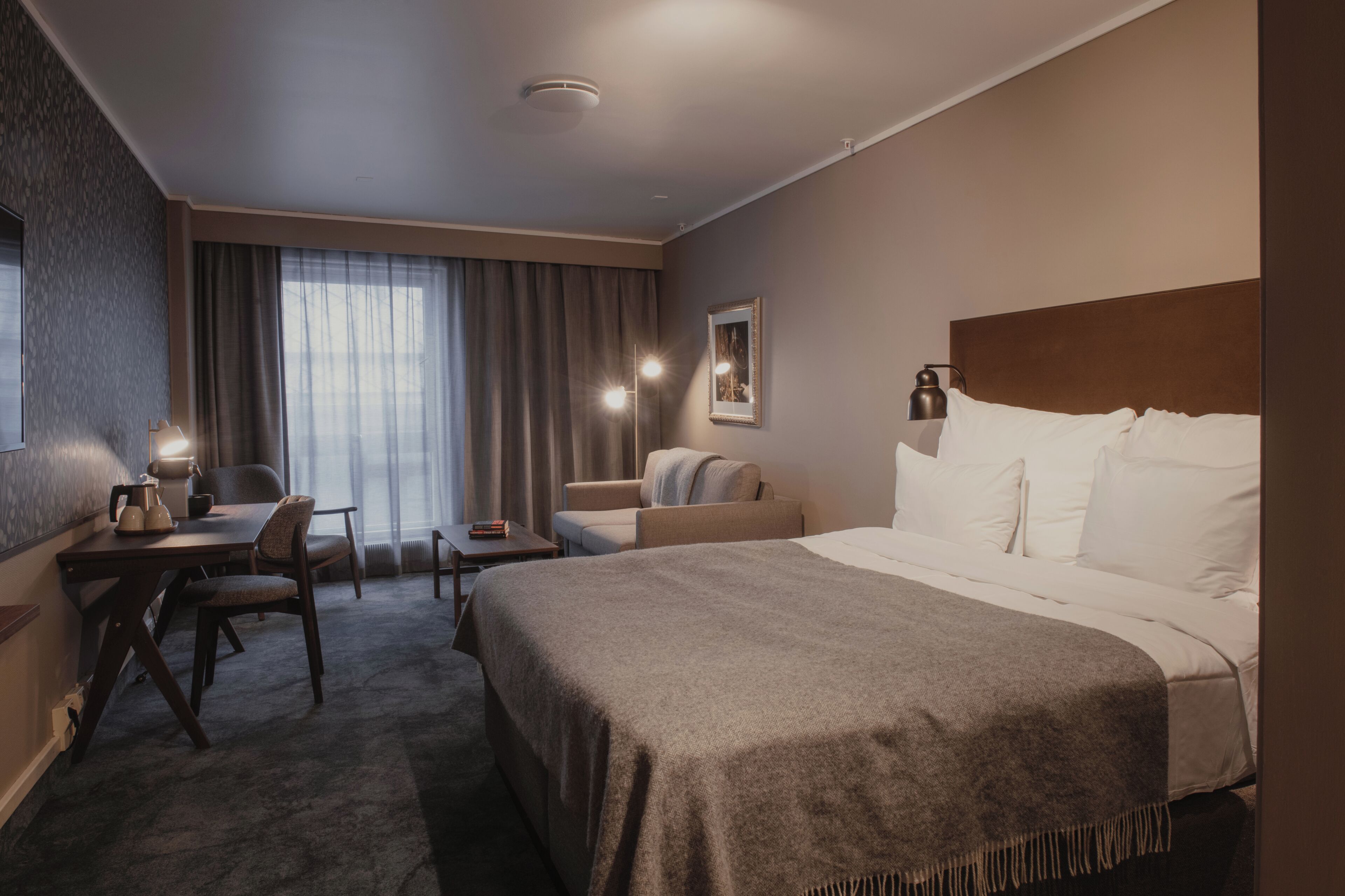 A hotel room with a bed, a desk, a chair, a sofa, and a window with curtains.