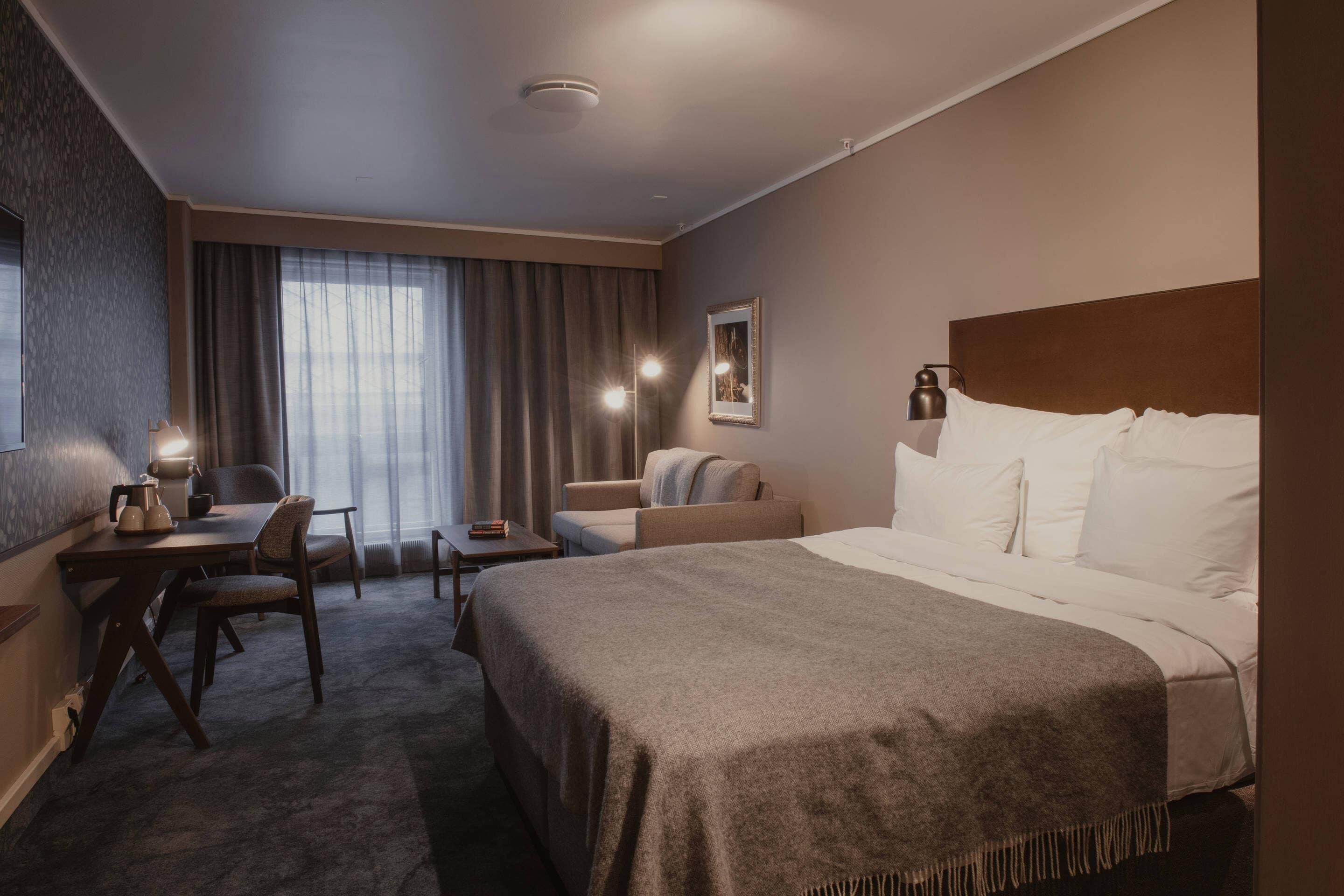 A hotel room with a bed, a desk with a chair, a sofa, and a window with curtains.