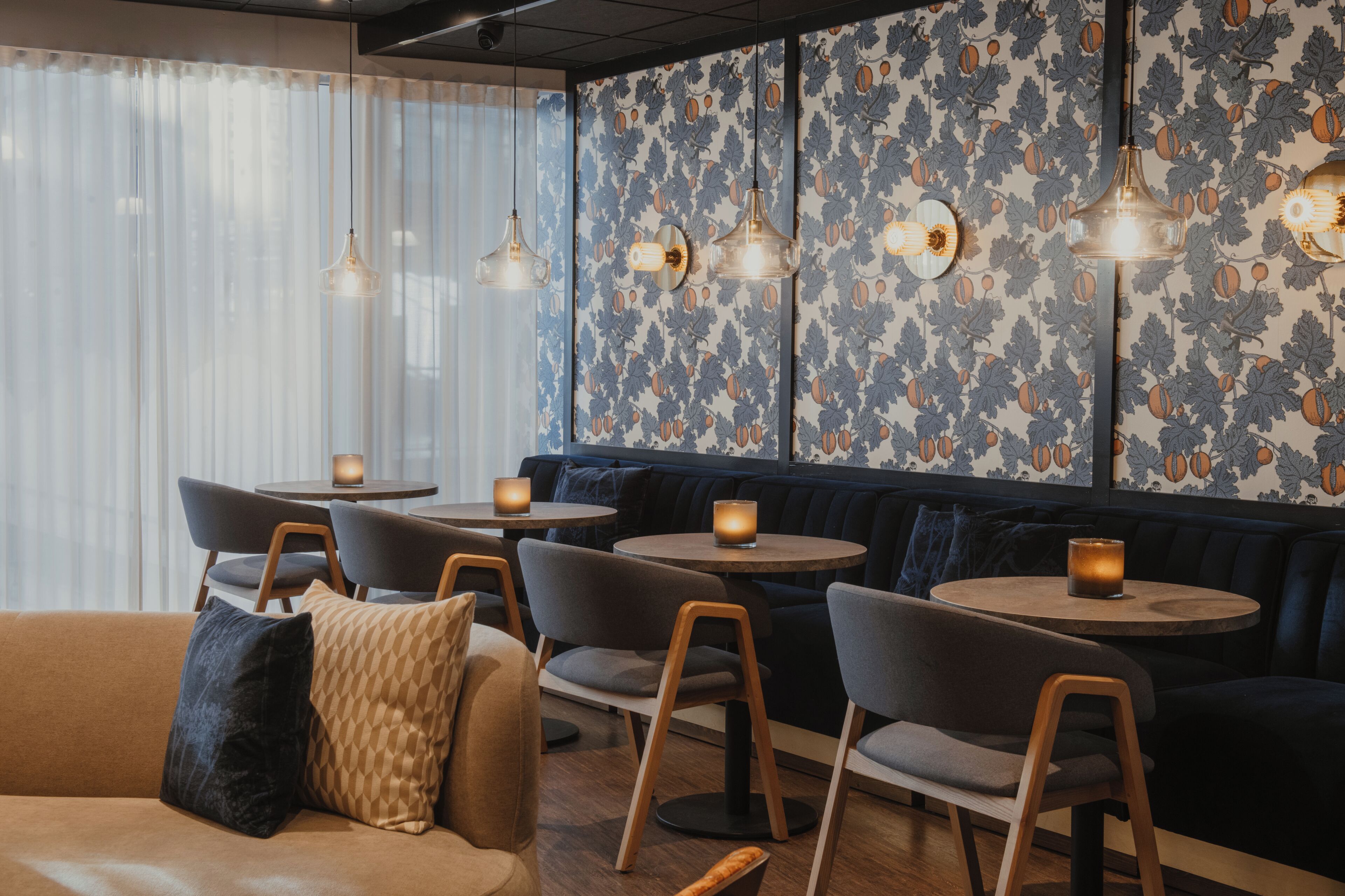 A restaurant interior with round tables, cushioned seating, pendant lighting, and patterned wallpaper.