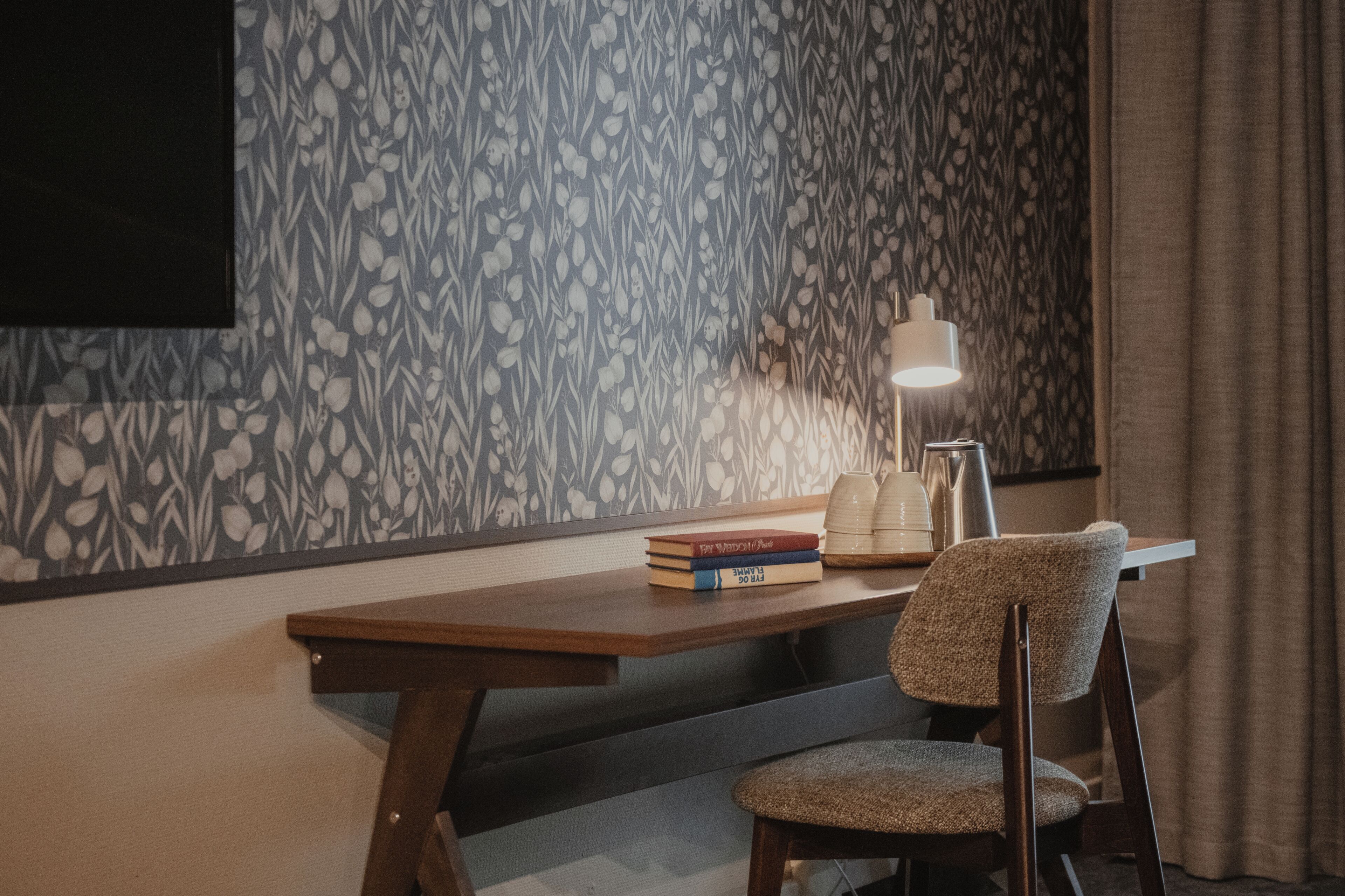 A room with a patterned wallpaper, a wooden desk, a chair, a lamp, and a few books.