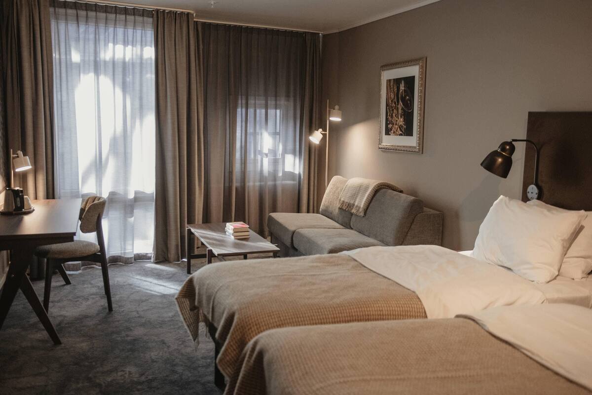 Hotellihuone, jossa on sänky, kaksi sohvaa, työpöytä, tuoli ja ikkuna, jossa on verhot.