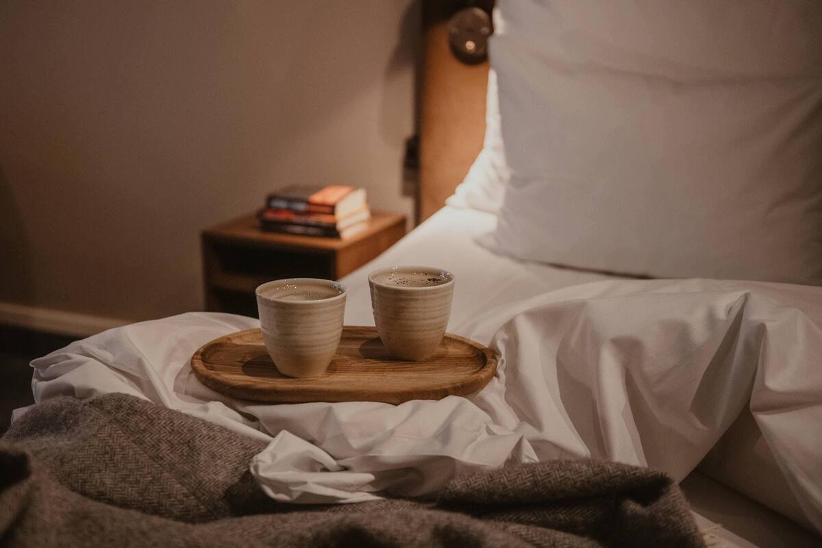 Una cama con ropa blanca, una mesita de noche de madera con libros y dos tazas de cerámica sobre una bandeja.