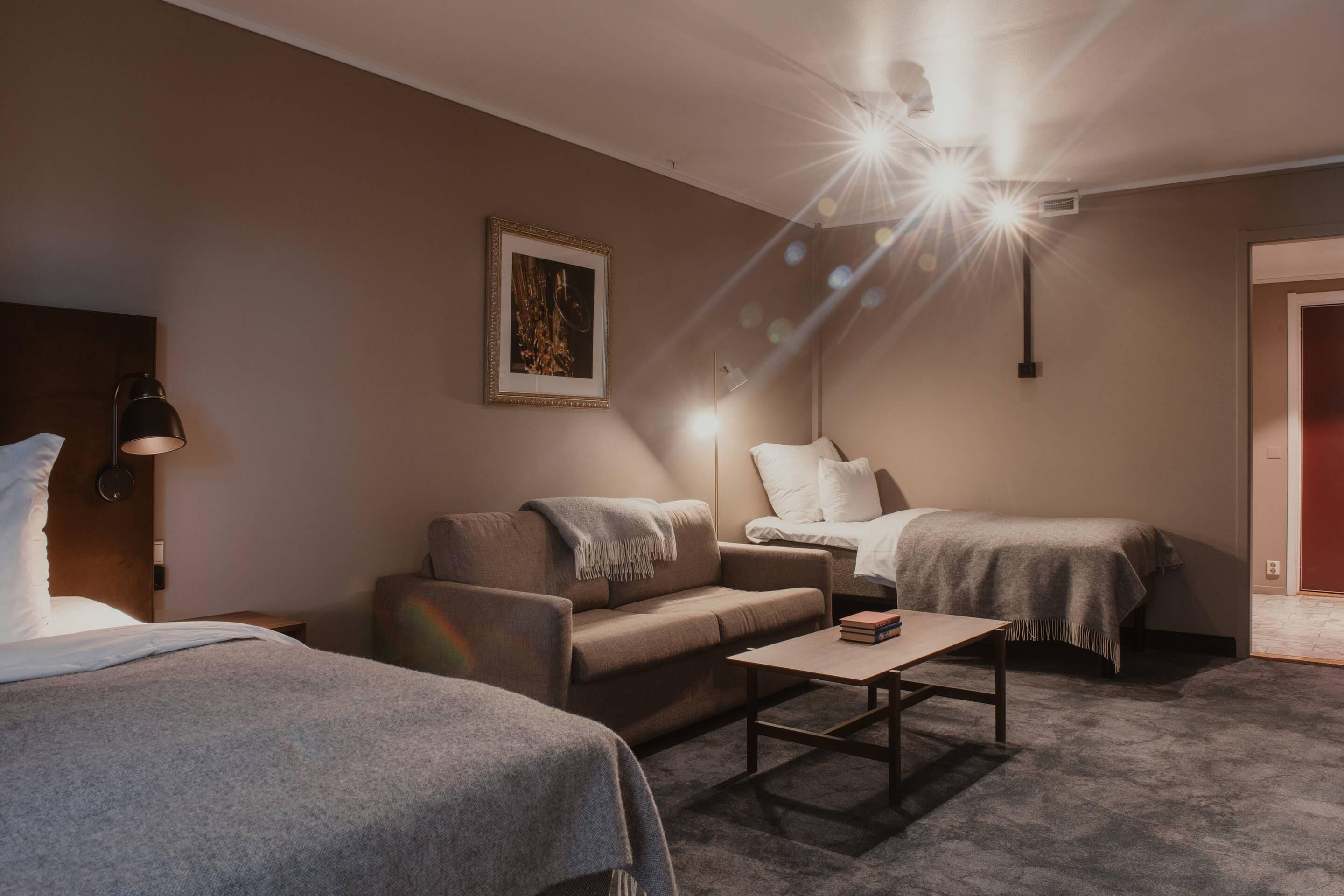 A hotel room with a sofa, two single beds, a small table, and a framed picture on the wall.