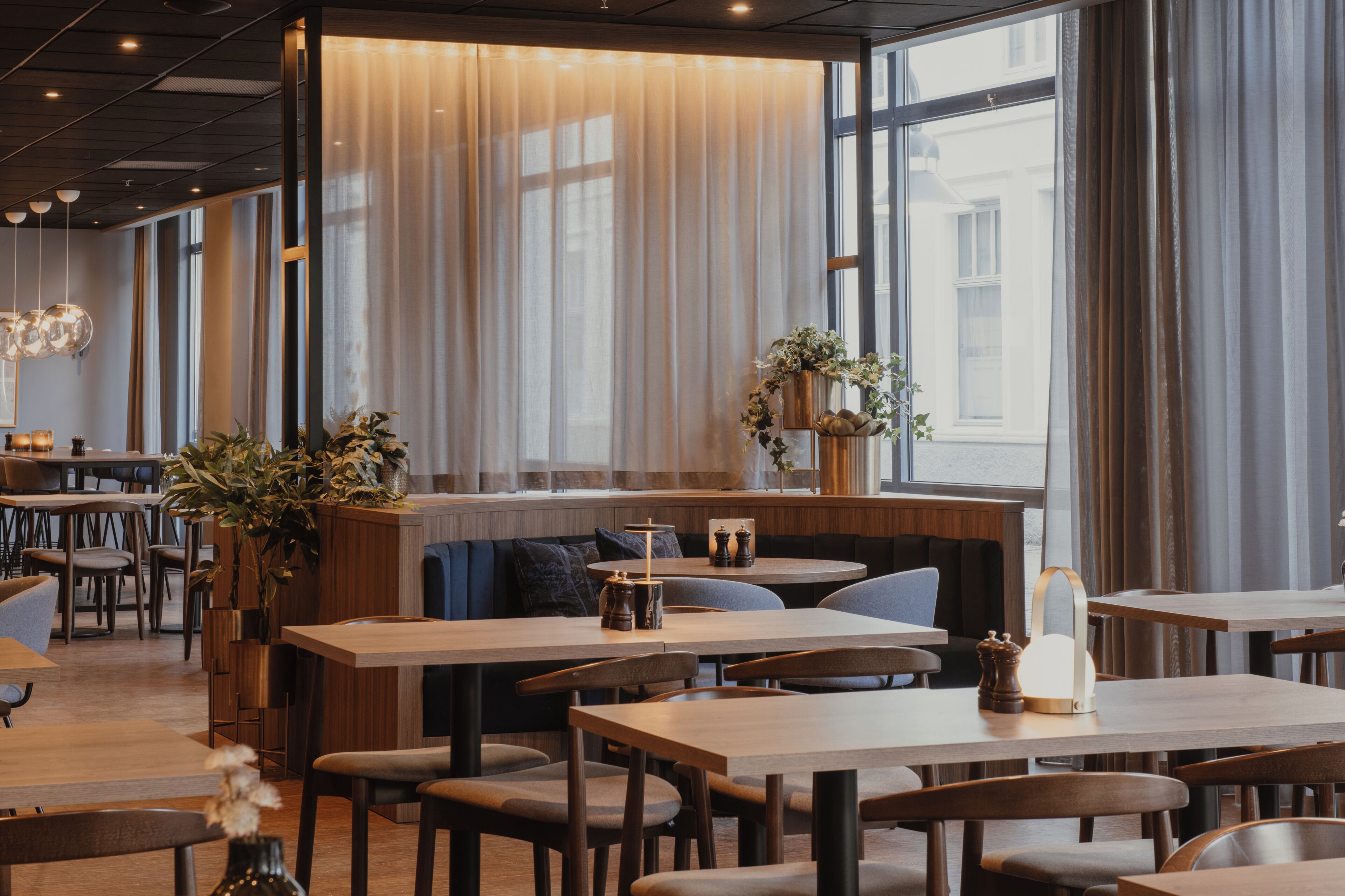 A restaurant with wooden tables and chairs, a seating area with cushions, and large windows with curtains.