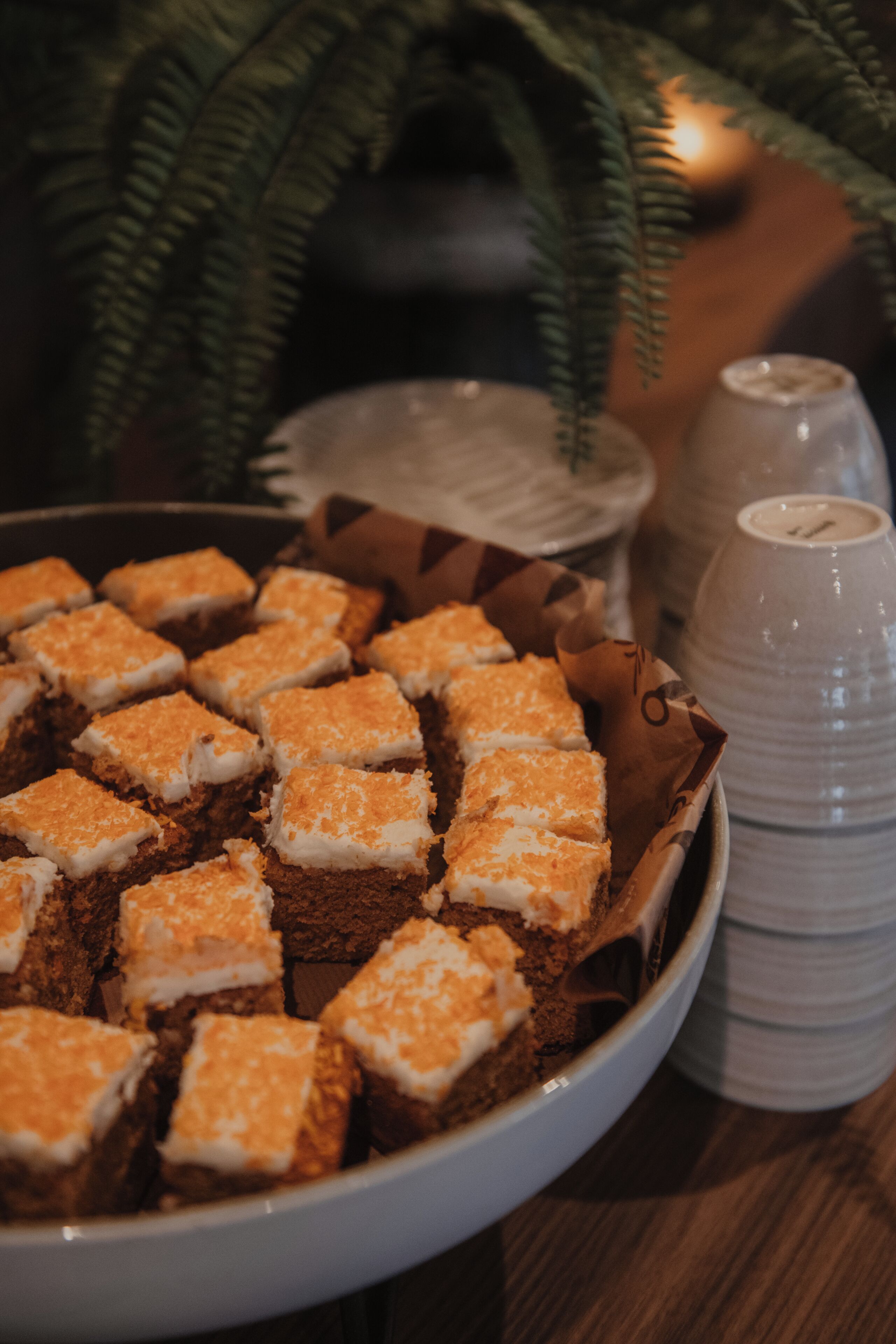 A tray of square-shaped desserts with a white topping, accompanied by stacked plastic cups and a salt shaker on a table.