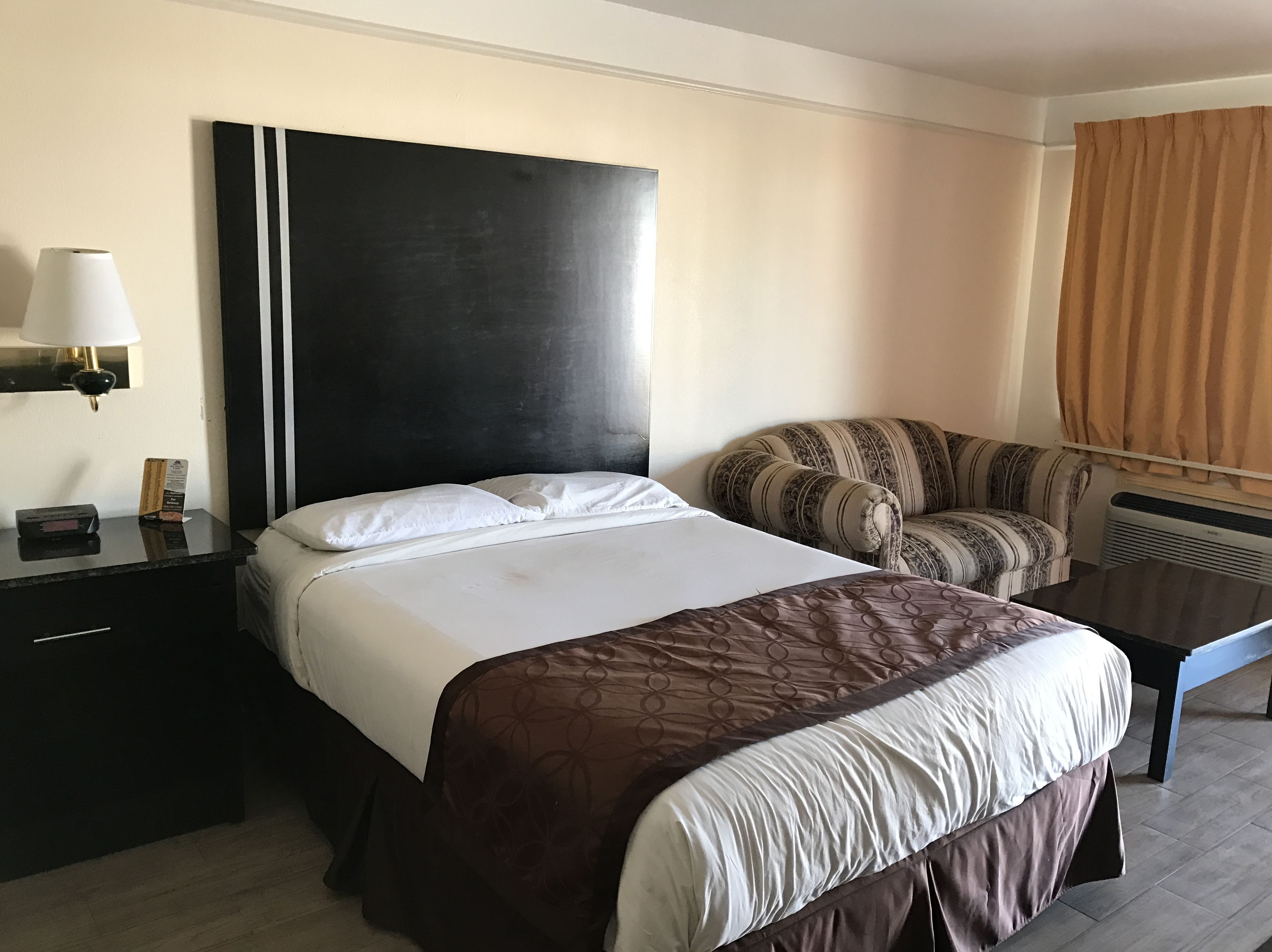 A hotel room with a large bed, a bedside table with a lamp, a chair, and a window with curtains.