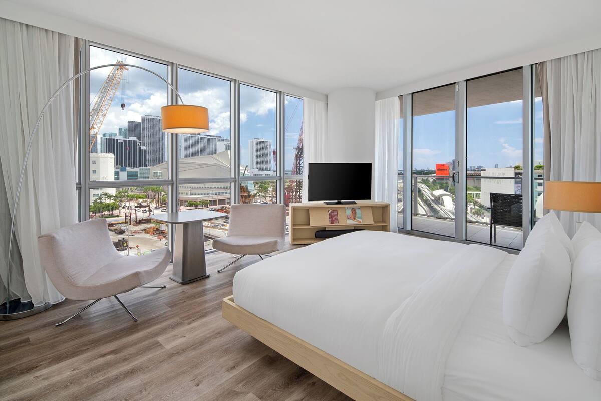Een moderne hotelkamer met een groot bed, een bureau, een stoel, een televisie en grote ramen met uitzicht op de stad.