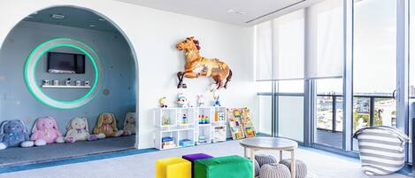 Une pièce spacieuse avec une grande fenêtre, un téléviseur fixé au mur, des jouets colorés et un espace de jeux avec des sièges confortables.
